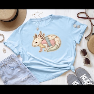 May include: Pale blue t-shirt with a patchwork axolotl design. The axolotl is made from pastel-coloured fabric pieces, including pink, blue, and yellow, with floral patterns. A casual, everyday wear item.
