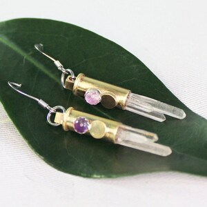 May include: A pair of gold earrings with clear quartz crystals and purple and pink gemstones. The earrings are hanging from a green leaf.