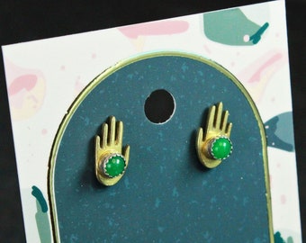 May include: A pair of gold earrings with green gemstone cabochons set in the centre of each earring. The earrings are shaped like hands with the fingers pointing upwards.