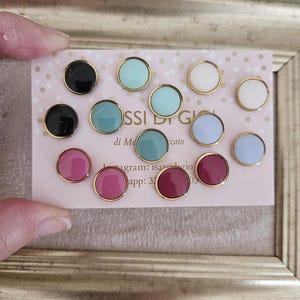 Può includere: Una collezione di orecchini a bottone rotondi in vari colori, tra cui nero, verde acqua, bianco, rosa e bordeaux. Ogni orecchino ha un bordo dorato ed è esposto su una scheda rosa con testo. Gli orecchini sono in una cornice dorata.