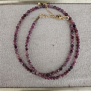 Può includere: Una delicata collana con piccole pietre preziose viola sfaccettate infilate insieme. La collana ha un design a doppio filo ed è assicurata con una chiusura color oro. Il gioiello è esposto su una superficie dai toni neutri.