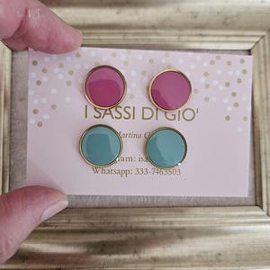 Può includere: Due paia di orecchini a bottone, uno rosa e uno turchese, con bordi dorati, sono esposti su una carta rosa. La carta presenta il testo "I Sassi Di Gio" e informazioni di contatto. Gli orecchini sono incorniciati in oro.