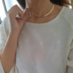 Può includere: Una collana a catena d'oro con un design delicato e leggermente strutturato. La collana è indossata da una persona con una camicia bianca.