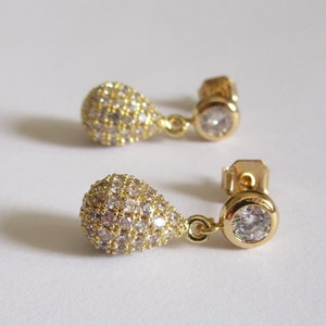 May include: A pair of gold-tone dangle earrings with clear cubic zirconia stones. The earrings feature a teardrop-shaped design with a smaller round stone at the top.