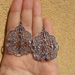 May include: A pair of silver filigree earrings with a delicate, intricate design. The earrings are shaped like a teardrop and have a floral pattern.