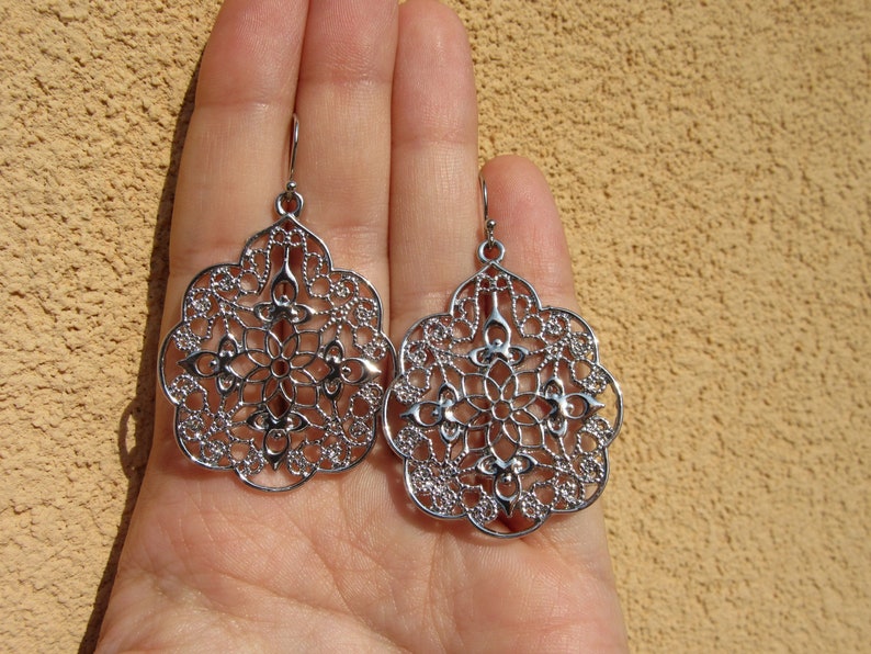 May include: A pair of silver filigree earrings with a delicate, intricate design. The earrings are shaped like a teardrop and have a floral pattern.