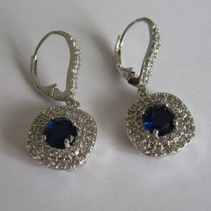 May include: A pair of silver dangle earrings with a blue oval gemstone surrounded by a halo of smaller clear gemstones.