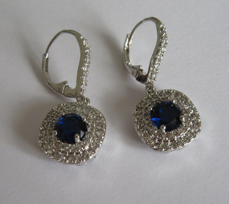 May include: A pair of silver dangle earrings with a blue oval gemstone surrounded by a halo of smaller clear gemstones.