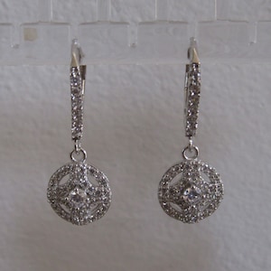 May include: A pair of silver dangle earrings with a round, crystal-encrusted design. The earrings have a drop-shaped design with a small, round crystal in the center of the larger round design.