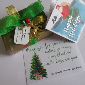 May include: A gold gift box wrapped in brown paper with a green and gold ribbon. A small gold reindeer charm hangs from the ribbon. A white card with the text "Thank you for your order. Wishing you a very merry Christmas and a happy new year." is next to the box. A smaller card with a watercolour illustration of Santa Claus and the text "Happy Holidays" is also visible.
