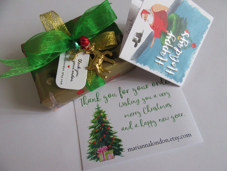 May include: A gold gift box wrapped in brown paper with a green and gold ribbon. A small gold reindeer charm hangs from the ribbon. A white card with the text "Thank you for your order. Wishing you a very merry Christmas and a happy new year." is next to the box. A smaller card with a watercolour illustration of Santa Claus and the text "Happy Holidays" is also visible.