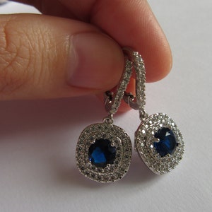 May include: Silver dangle earrings with a blue gemstone surrounded by a halo of smaller clear gemstones.