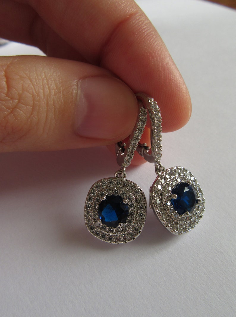 May include: Silver dangle earrings with a blue gemstone surrounded by a halo of smaller clear gemstones.