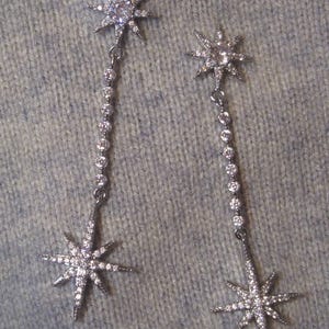Puede incluir: Un par de pendientes plateados con forma de estrella. Cada pendiente presenta una estrella en la parte superior e inferior, conectadas por una cadena de pequeñas piedras redondas y transparentes. Las estrellas están incrustadas con pequeñas piedras claras, creando un efecto brillante.