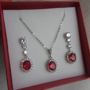 Peut inclure: Un collier et des boucles d'oreilles en argent avec une pierre ovale rouge entourée de petites pierres claires. Les bijoux sont présentés dans une boîte rouge.