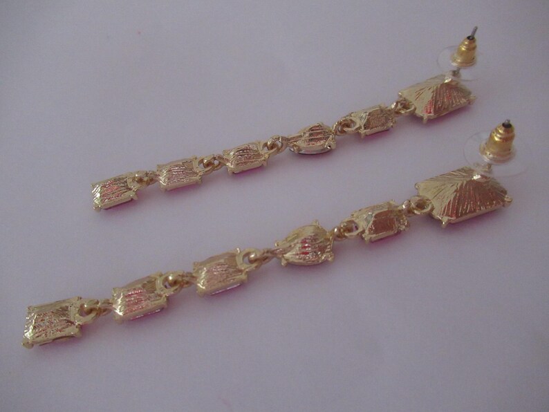 May include: A pair of long, gold earrings with pink rectangular stones. The earrings have a dangling design with multiple stones.