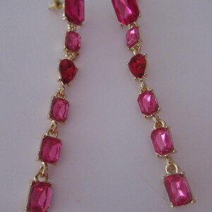 May include: A pair of long, dangling earrings with pink and red faceted gemstones. The earrings are made of gold-toned metal and have a simple, elegant design.