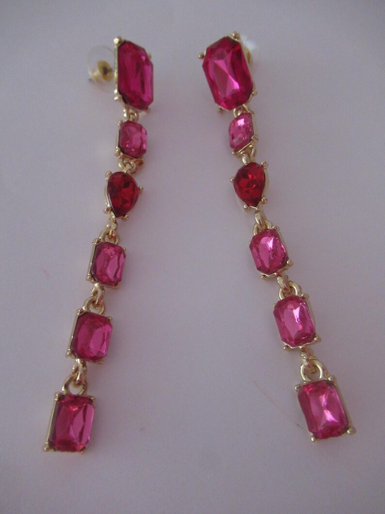 May include: A pair of long, dangling earrings with pink and red faceted gemstones. The earrings are made of gold-toned metal and have a simple, elegant design.