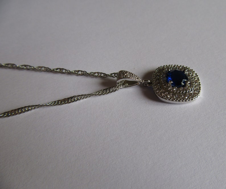 May include: A silver chain necklace with a pendant featuring a large blue gemstone surrounded by smaller clear gemstones.