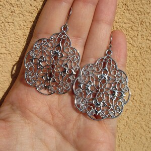 May include: A pair of silver filigree earrings with a delicate, intricate design. The earrings are shaped like a teardrop and have a floral pattern.