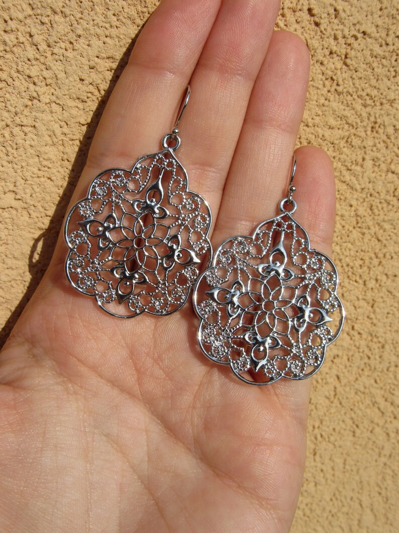 May include: A pair of silver filigree earrings with a delicate, intricate design. The earrings are shaped like a teardrop and have a floral pattern.