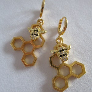 Puede incluir: Pendientes de tono dorado con un diseño de panal y un pequeño dije de abeja con detalles en blanco y negro.