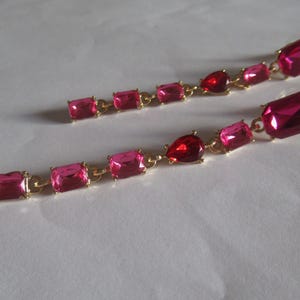 May include: A pair of long pink and red crystal earrings with gold chain links. The earrings feature rectangular and teardrop shaped crystals.