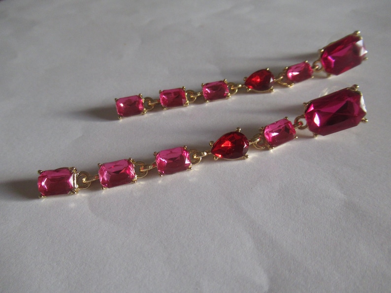 May include: A pair of long pink and red crystal earrings with gold chain links. The earrings feature rectangular and teardrop shaped crystals.