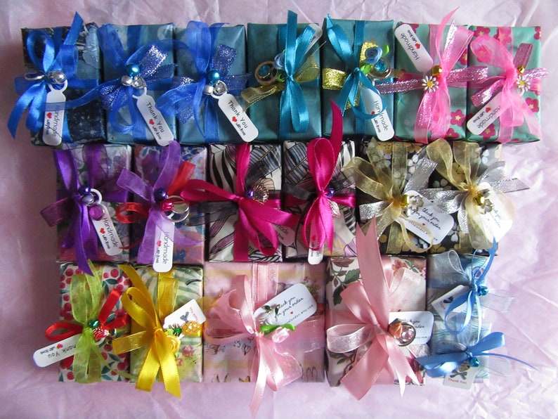 May include: A collection of small, wrapped gift boxes with various colorful patterned paper, ribbons, and bows. The boxes are tied with different colored ribbons, including blue, pink, yellow, purple, and green. Some boxes have small charms or embellishments attached to them. The boxes are arranged in a grid pattern on a pink surface. The boxes are all approximately the same size and shape.