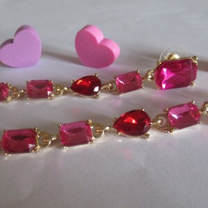 May include: A pair of long, dangling earrings with pink and red gemstones. The earrings are made of gold metal and have a teardrop-shaped gemstone in the center.