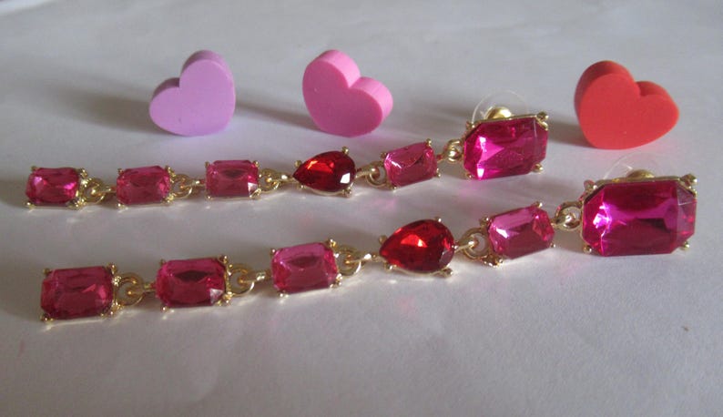 May include: A pair of long, dangling earrings with pink and red gemstones. The earrings are made of gold metal and have a teardrop-shaped gemstone in the center.