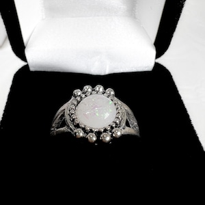 Puede incluir: Un anillo plateado con una piedra blanca iridiscente grande, engastada en un bisel con cuentas. El anillo se muestra en una caja de joyería negra con un interior de satén blanco, mostrando el diseño y el color del anillo.