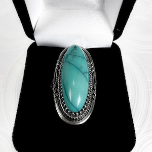 Puede incluir: Anillo plateado con una gran piedra turquesa ovalada con vetas oscuras. La piedra está engastada en un marco decorativo y texturizado. El anillo se presenta en una caja de joyería negra con forro de satén blanco.