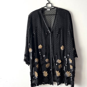 Può includere: Una giacca nera in tessuto trasparente con paillettes dorate e argentate a forma di foglie. La giacca ha un collo a V e una chiusura con bottone.