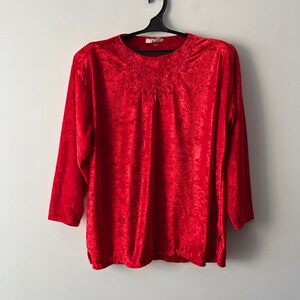 Könnte beinhalten: Ein langärmliges rotes Samt-Shirt mit Rundhalsausschnitt und einem kleinen Stickmuster auf der Vorderseite. Das Shirt hängt an einem schwarzen Kleiderbügel an einer weißen Wand.