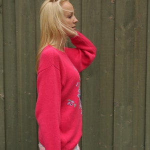 May include: A woman wearing a pink sweater with floral embroidery and floral print pants. She is standing in front of a wooden wall.