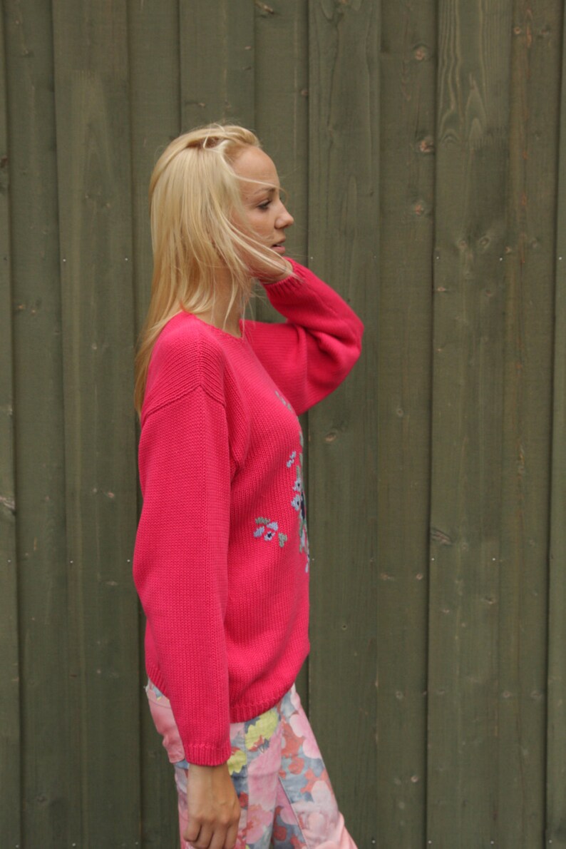 May include: A woman wearing a pink sweater with floral embroidery and floral print pants. She is standing in front of a wooden wall.