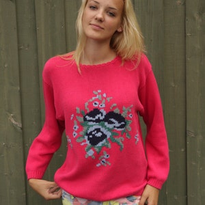 May include: A pink knitted sweater with a floral design in shades of green, black, and white. The sweater has a crew neckline and long sleeves.
