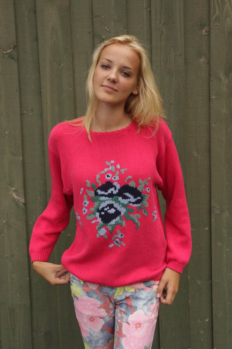 May include: A pink knitted sweater with a floral design in shades of green, black, and white. The sweater has a crew neckline and long sleeves.