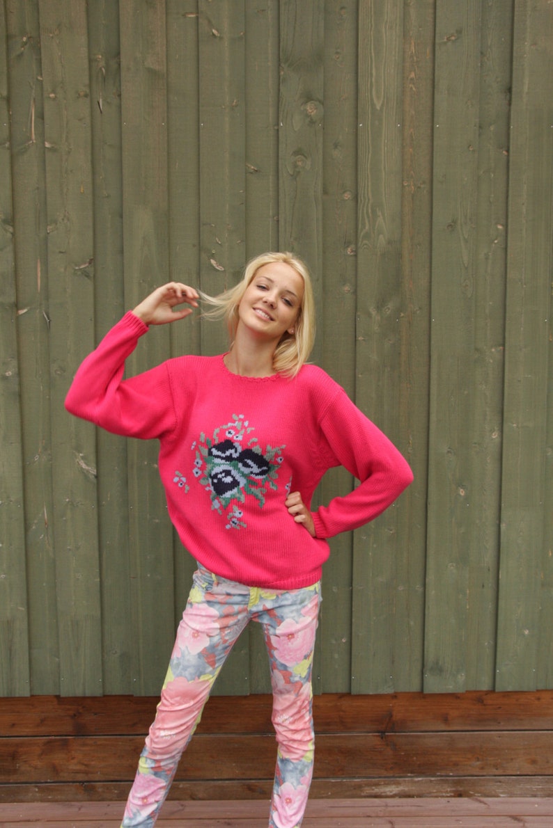 May include: A woman wearing a pink sweater with a floral design and floral print pants. She is standing in front of a green wooden wall.