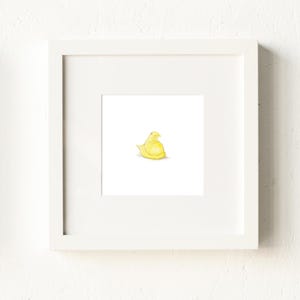 May include: A framed watercolor print featuring a yellow chick. The artwork is set against a white background and framed in a white square frame. The image is mounted on a white wall.