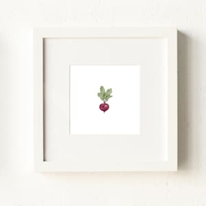 May include: A framed watercolor print featuring a single beet. The beet is a deep red-purple color with green leaves. The artwork is set against a white background and framed in a white square frame.