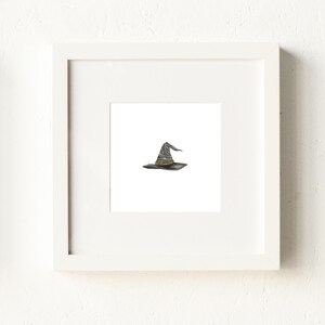 May include: A framed print featuring a watercolor illustration of a witch's hat. The hat is primarily gray with a slightly curved brim and a pointed crown. The artwork is set against a white background within a white frame.