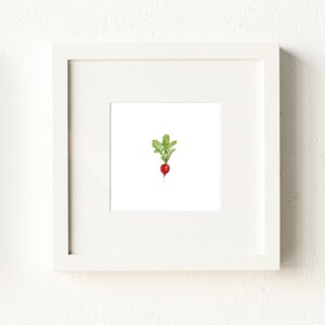 May include: A framed art print featuring a watercolor illustration of a radish. The radish is red with green leaves, set against a white background. The artwork is displayed in a white square frame, suitable for kitchen or dining room decor.