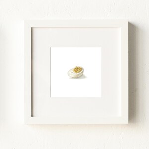 May include: A framed watercolor print of a halved deviled egg. The egg white is a creamy white, and the yolk filling is a mix of yellow and brown. The artwork is set against a white background and framed in a white square frame.