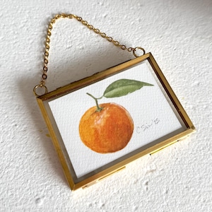 May include: A framed watercolor painting of an orange fruit with a green leaf. The artwork is in a gold-colored rectangular frame with a gold chain for hanging. The painting is signed "C. Sain '15".