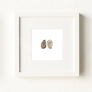 May include: A framed watercolor print featuring two oyster shells. One shell is closed, displaying a textured brown exterior, while the other is open, revealing a pearlescent interior. The artwork is set against a white background within a white frame.