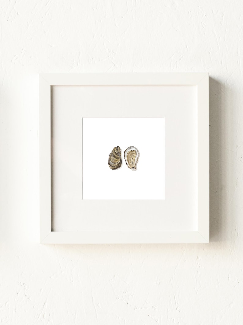 May include: A framed watercolor print featuring two oyster shells. One shell is closed, displaying a textured brown exterior, while the other is open, revealing a pearlescent interior. The artwork is set against a white background within a white frame.