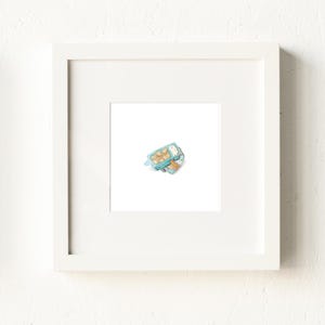 May include: A framed watercolor print featuring a light blue lunchbox filled with pastries and a small orange. The artwork is set against a white background and framed in a white wooden frame.