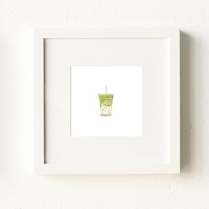 May include: A framed watercolor print of a matcha latte in a clear plastic cup with a straw. The drink is a light green color, and the cup has a white lid. The frame is white.